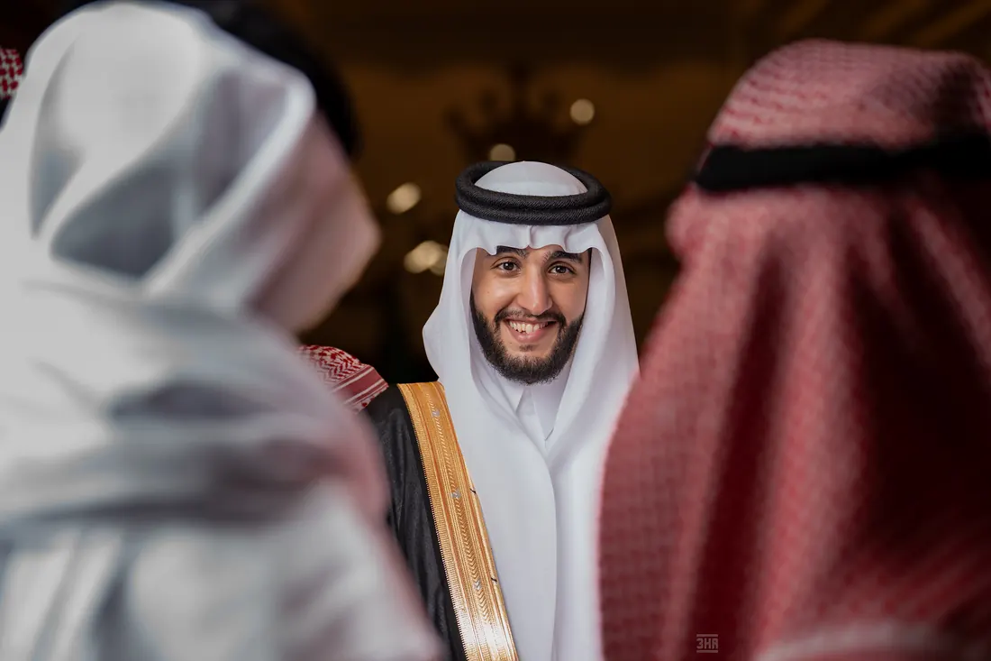 عريس مبتسم يظهر بين كتفَي مهنئَين بالشماغ الأبيض والأحمر