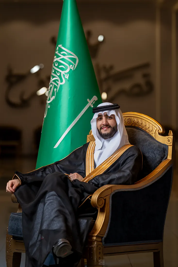 عريس جالس على كرسي مذهّب بجانب العلم السعودي