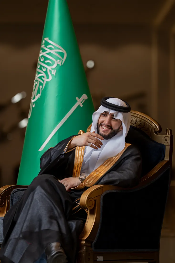 عريس يضحك جالسًا على كرسي مذهّب بجانب العلم السعودي