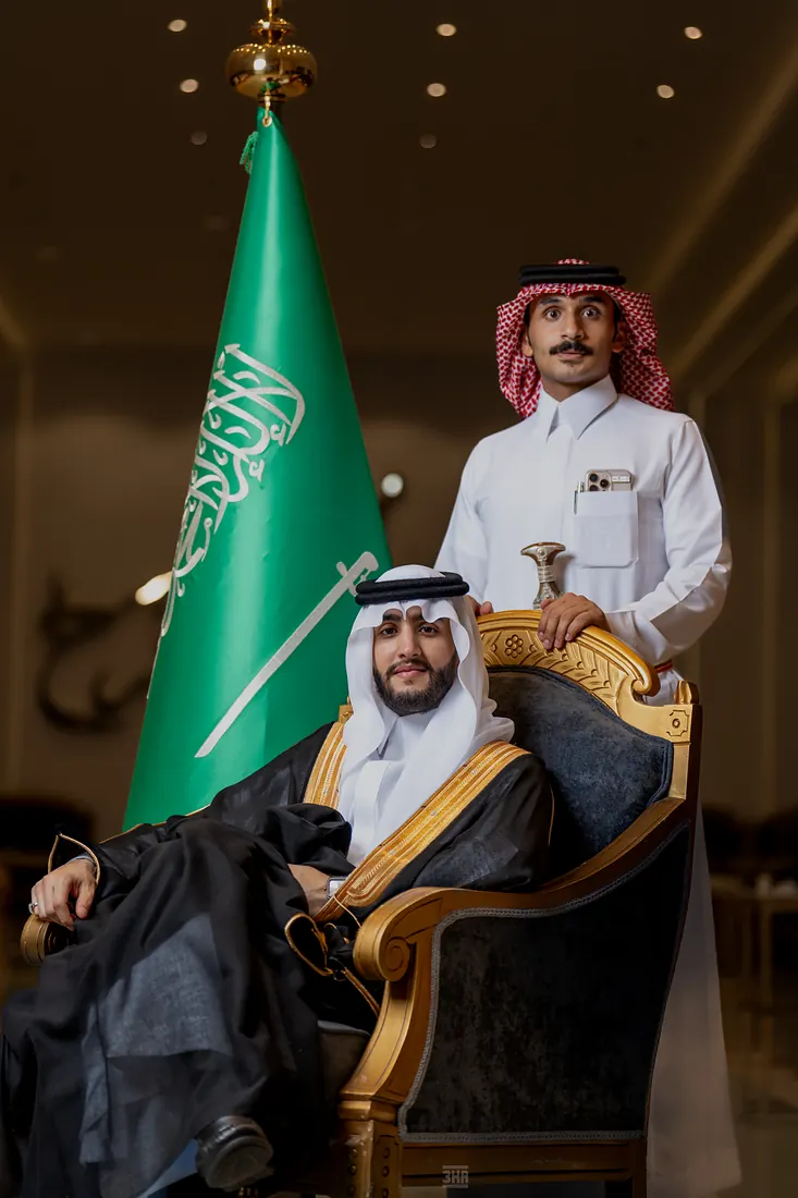 عريس جالس على كرسي مذهّب ورجل بالشماغ الأحمر يقف خلفه بجانب العلم السعودي