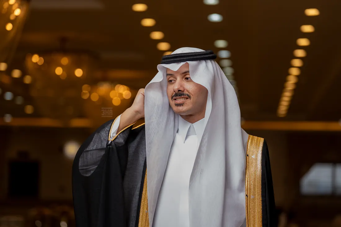 عريس يعدّل شماغه الأبيض بيده في قاعة مضاءة بالثريات