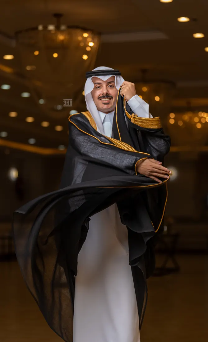 عريس يلوّح بطرف بشته الأسود المطرّز بالذهب تحت أضواء القاعة