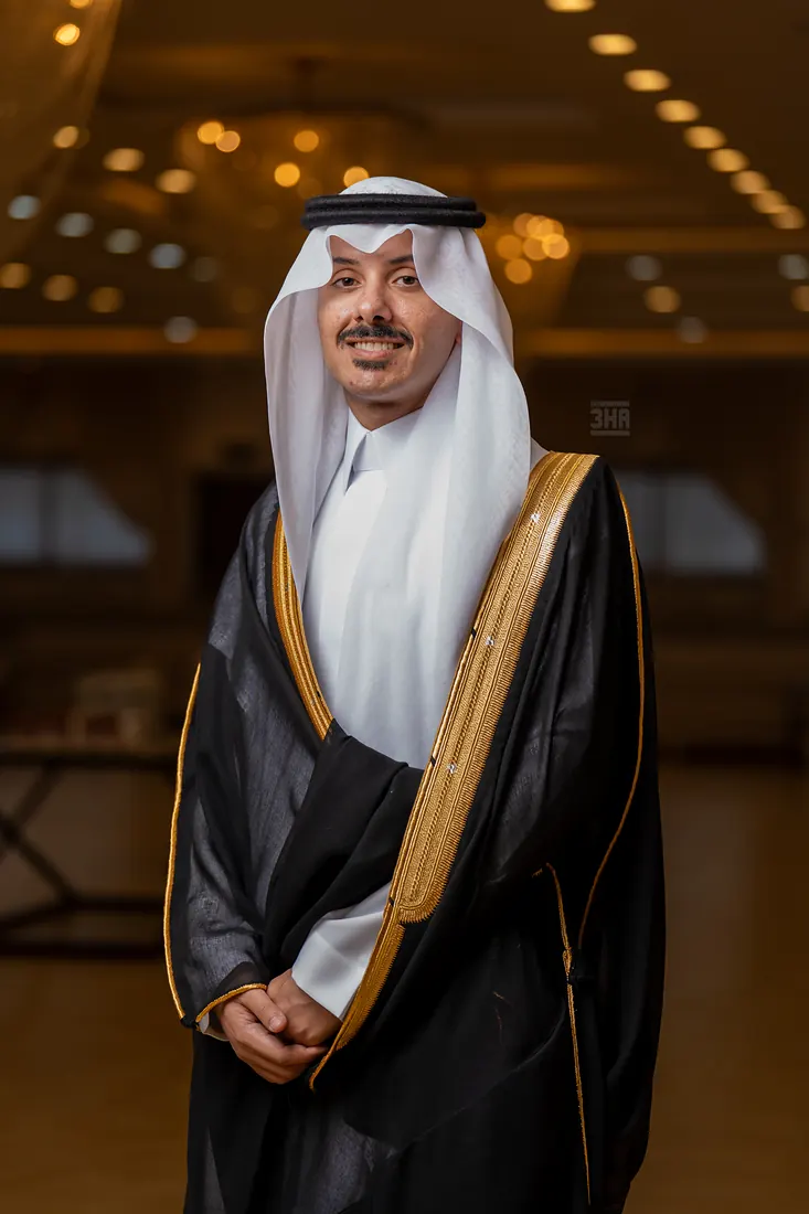 عريس بالبشت يقف مبتسمًا تحت أضواء القاعة