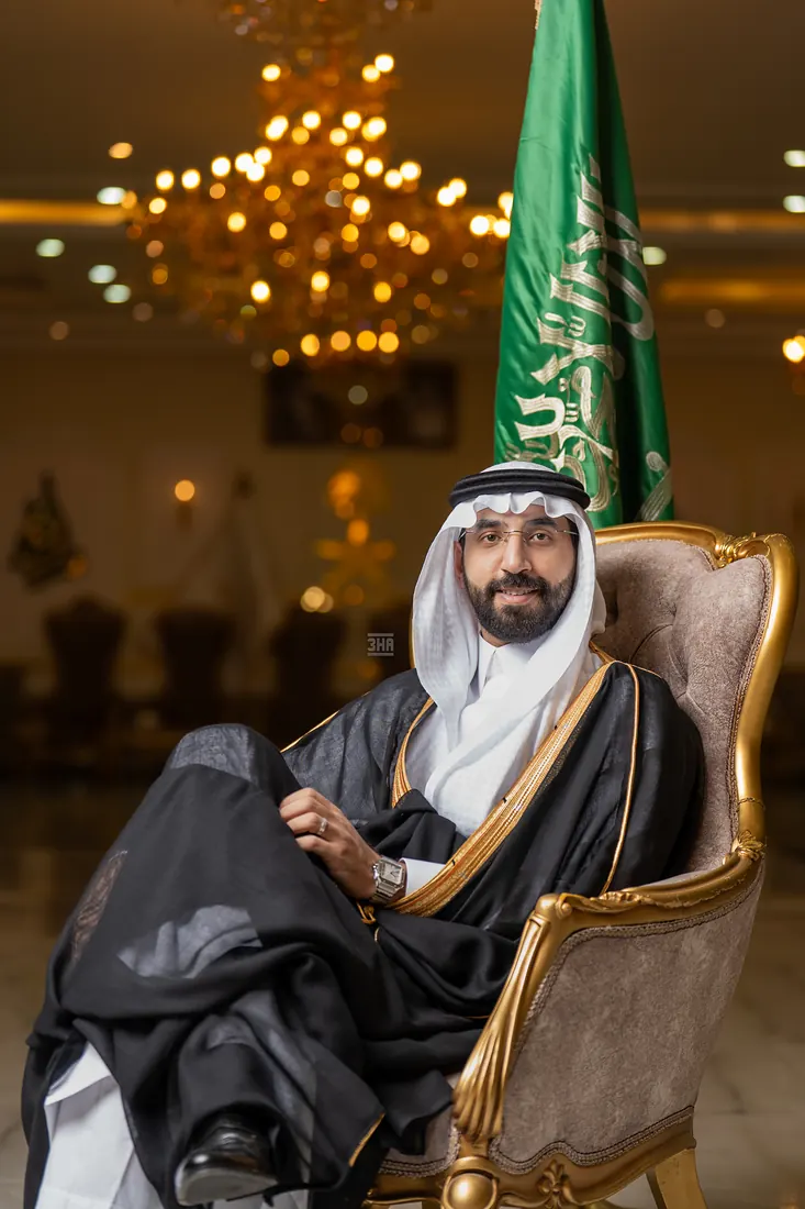 عريس بالبشت يجلس على كرسي مخملي واضعًا ساقًا على ساق أمام العلم السعودي